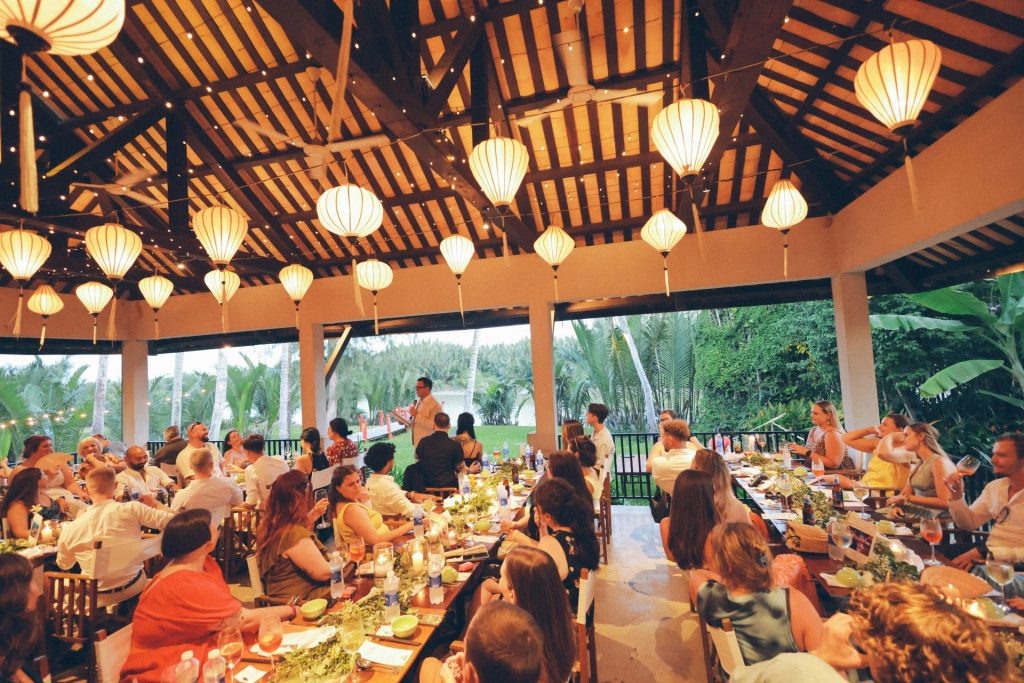 Menschen speisen an langen Tischen unter Laternen.